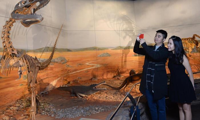 a young man and woman take a photo of a dinosaur skeleton. the skeleton is much taller than them and it shows sharp teeth. the picture was taken in th